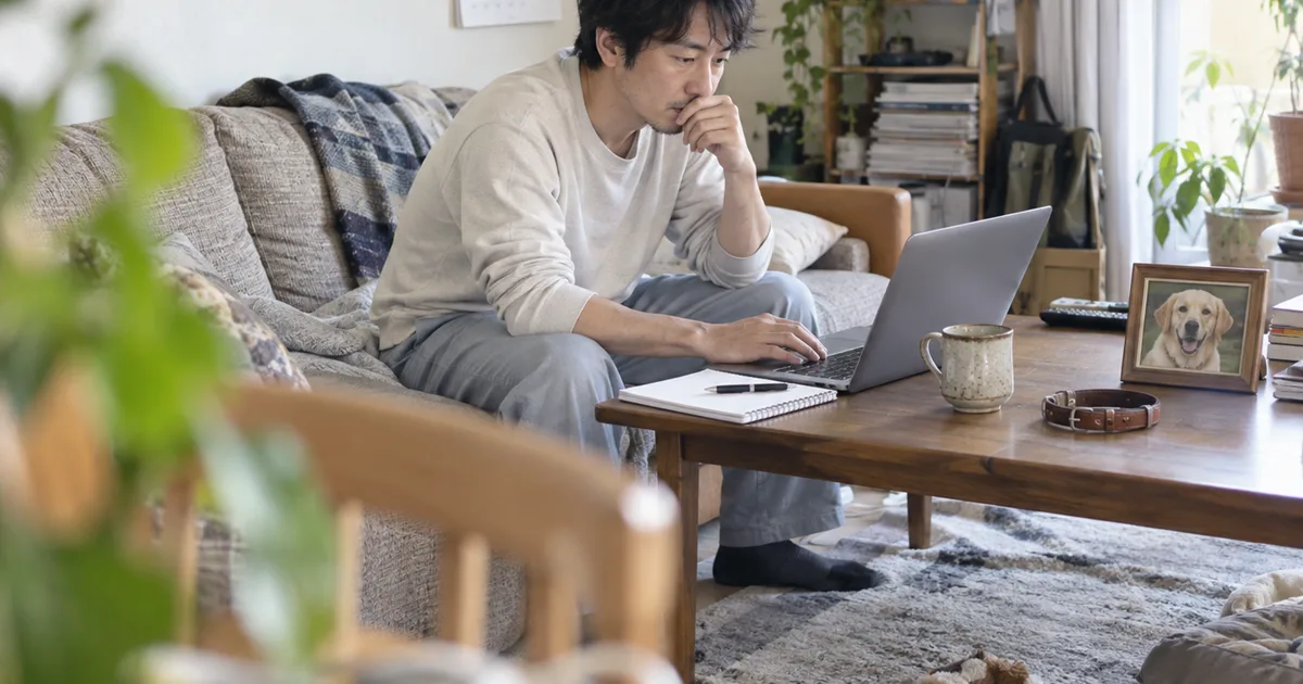 ソファでノートパソコンを見て調べる30代の男性と、机の上のラブラドールレトリバーの写真と首輪